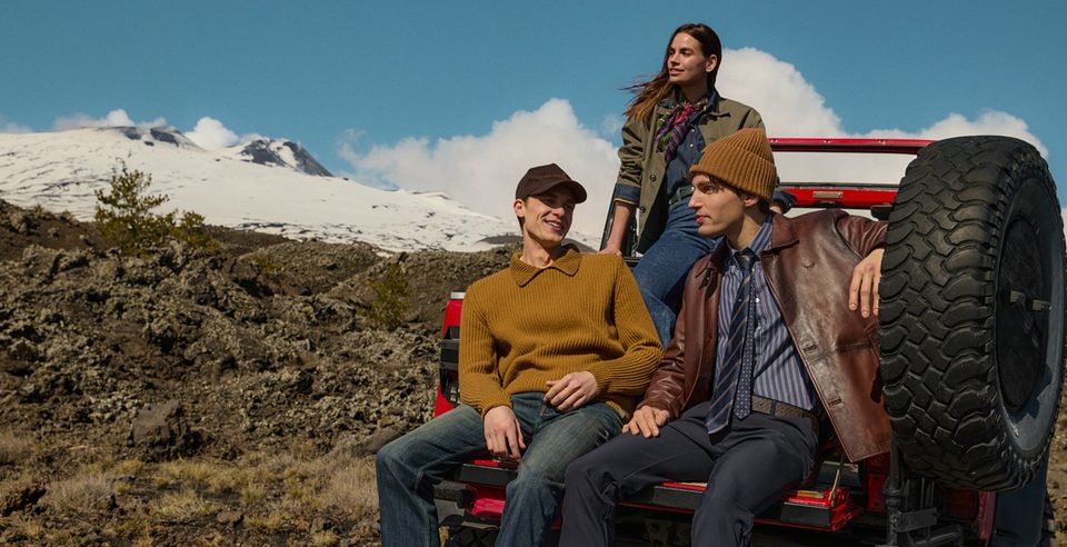 Tre modelli seduti sul retro di un fuoristrada rosso, con la colata lavica e l'Etna innevato alle spalle
